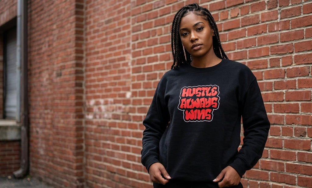 A woman with braided hair stands against a red brick wall wearing a black crewneck sweatshirt with a red and white graphic text that reads 'HUSTLE. EXIST.